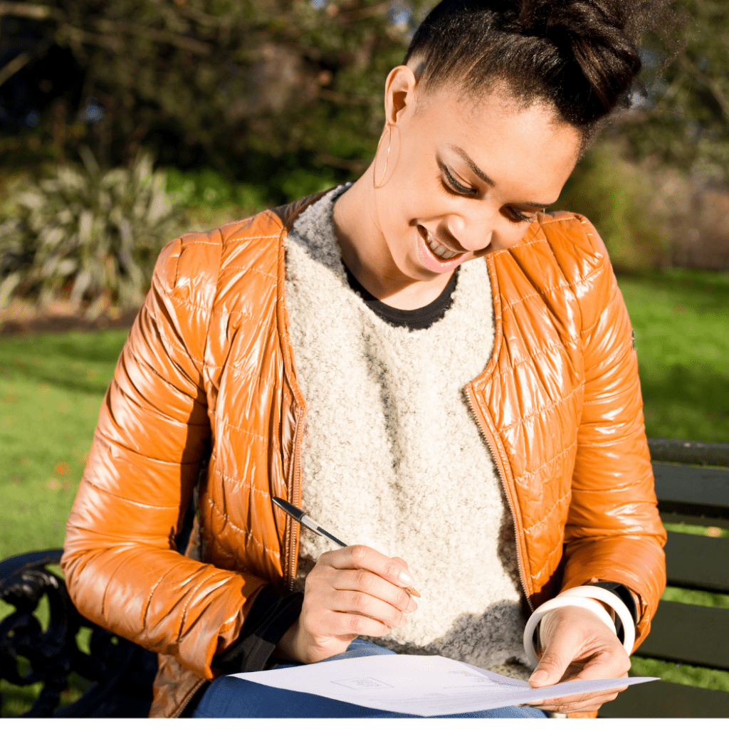 Happy women sitting on a park bench in an orange leather jacket and white sweater holding a pen and looking at a piece of paper on her lap