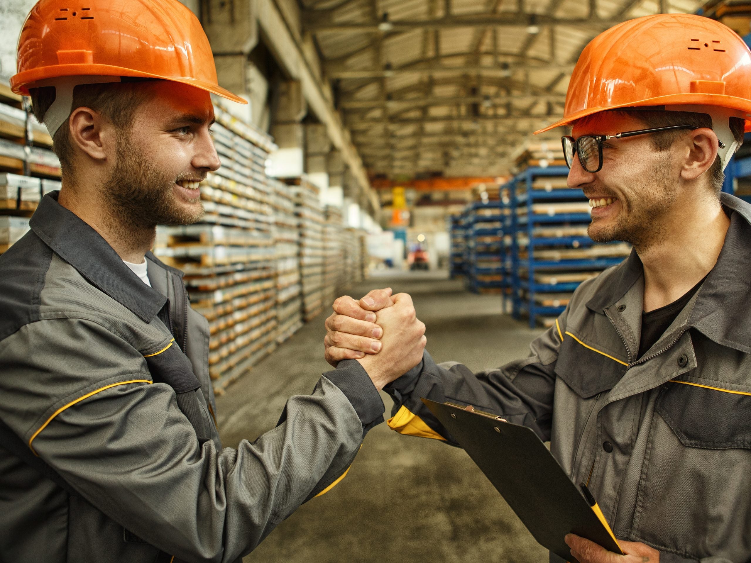 Shot of two cheerful male workers in hardhat and protective uniform shaking hands while working at the hardware factory manufacturing production plant positivity success teamwork collaboration