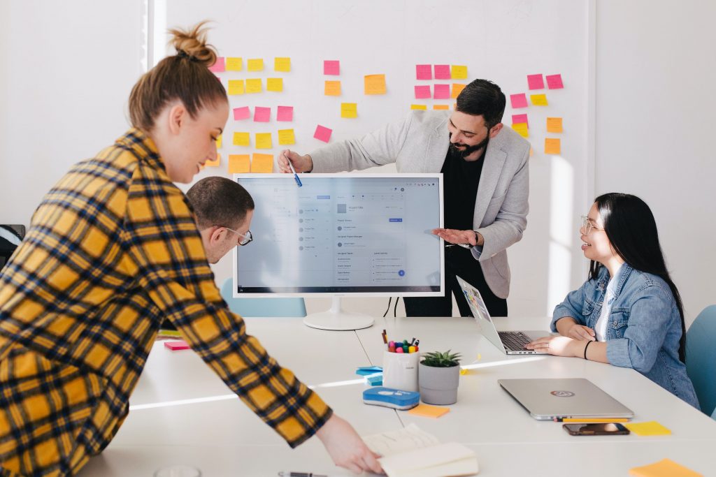 4 people at a conference room table as a man presents at a monitor with a wall of colorful sticky notes behind him