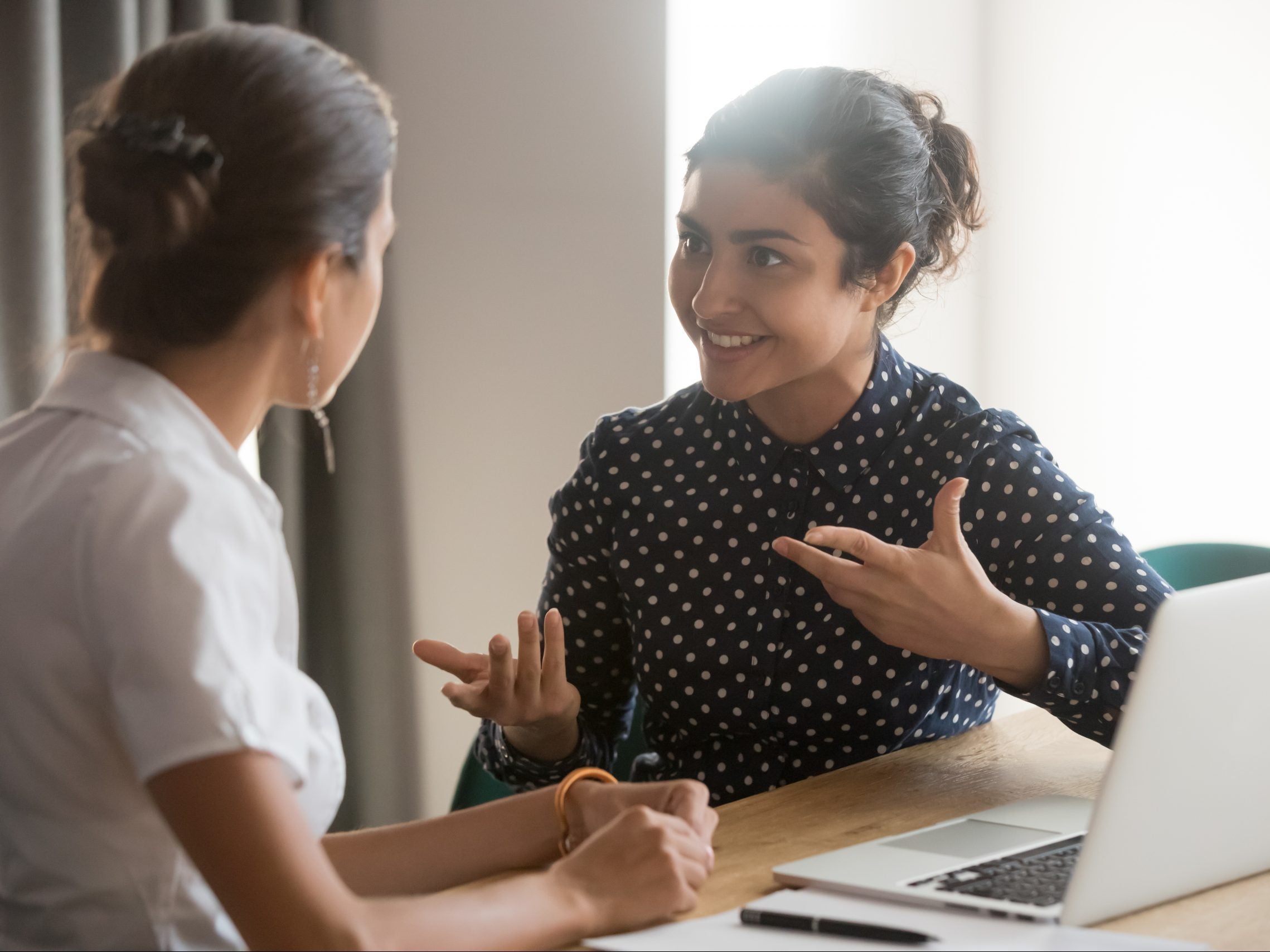 Excited,Multiethnic,Female,Employees,Discuss,Work,Issues,Sitting,At,Office Two women having a conversation while sitting at a table with a laptop