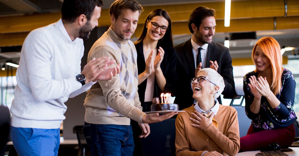 Group of people at woman's desk celebrating birthday with cake