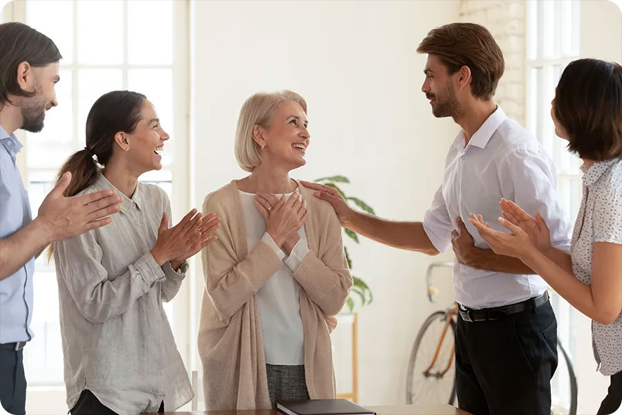 An older woman stands in the center of a group of people clapping, with one man putting his arm on the older lady, showing support and celebrating her.