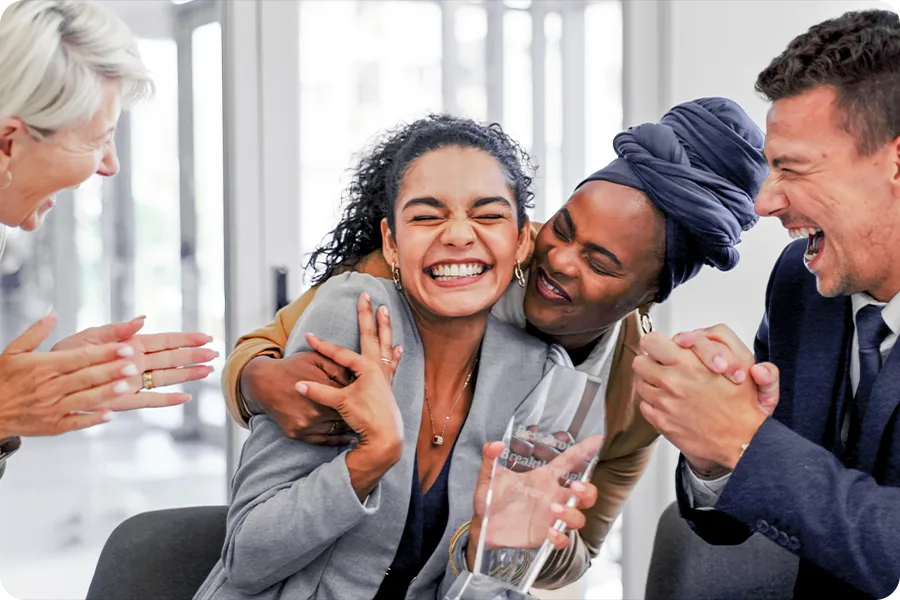 Two people are clapping, while another is holding the shoulders of a woman in the center of the frame. The woman is smiling brightly and holding a glass award.
