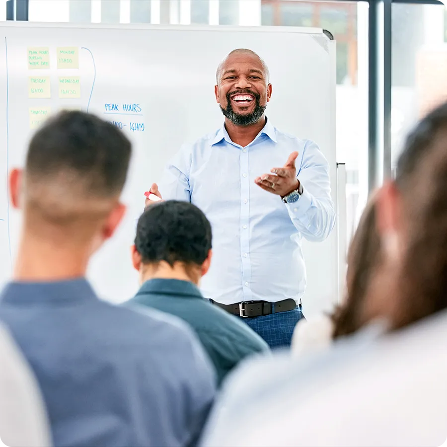 A man smiles at the front of a classroom of people, with a white board behind him.