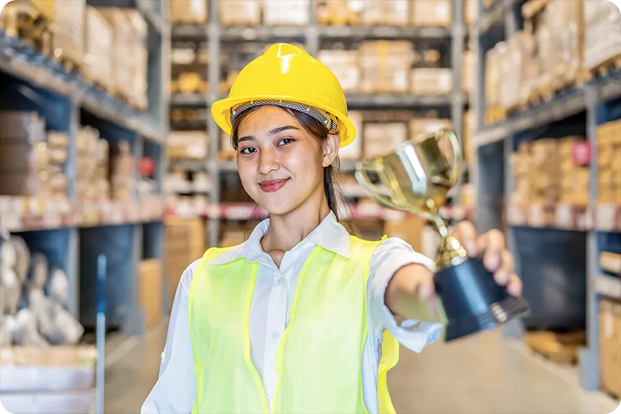A woman in a yellow hard hat is standing in a large warehouse and holding a trophy towards the camera. 
