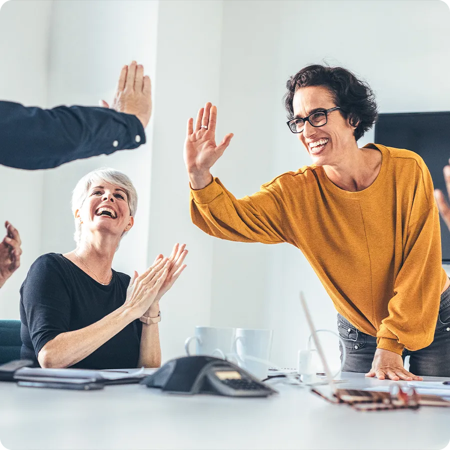 A woman in a mustard sweater is shown trying to high-five another person off camera. She is standing and leaning over a conference table with two other people visibly clapping nearby.