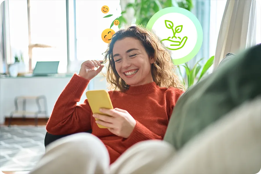 A woman is lounging on a couch in an auburn sweater and smiling at her yellow smart phone. Behind her is an icon signifying green energy and several smiling face emojis.