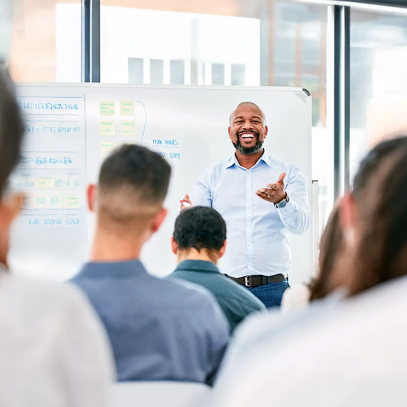 A man smiles at the front of a classroom of people, with a white board behind him.