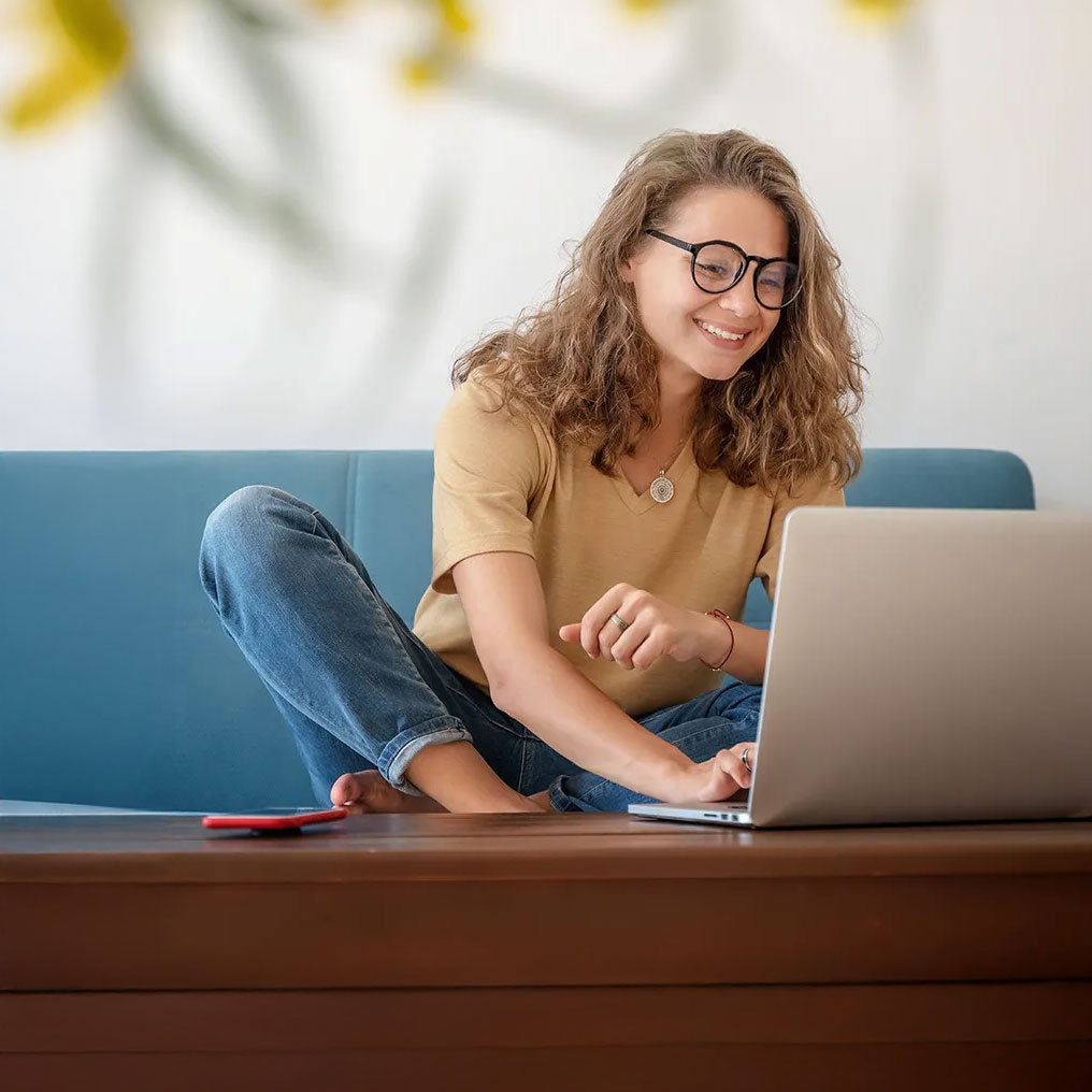 A woman in glasses and business casual attire sits cross-legged on a couch while working on her laptop.