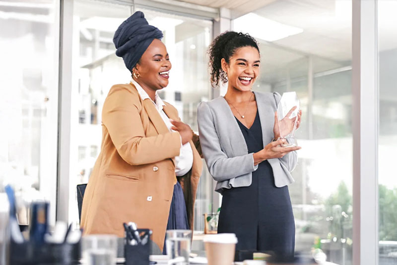 Two female employees smile while holding an award recognizing their efforts in a conference room.